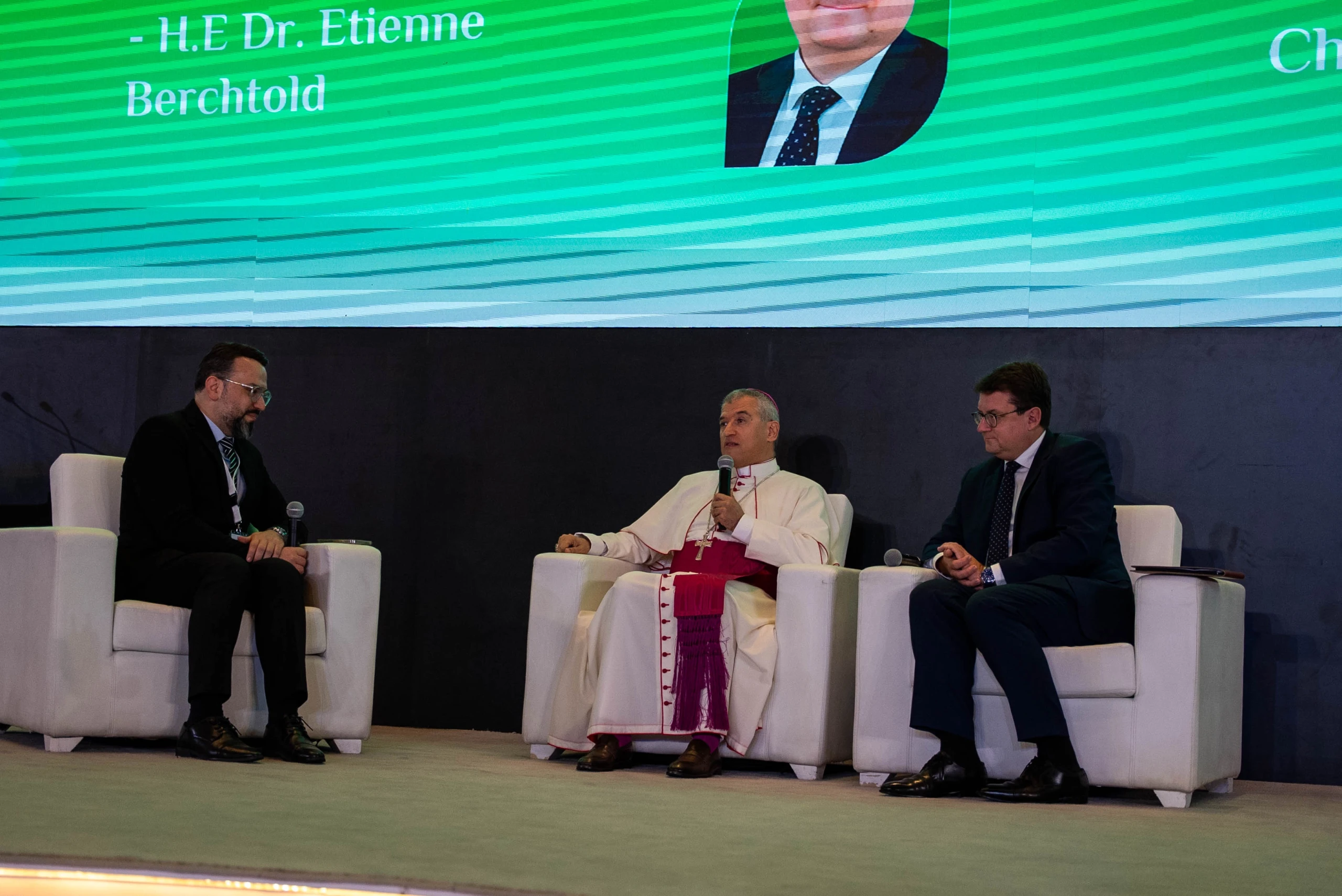 A clergyman in a white cassock with a fuchsia sash speaks on a panel beside two suited men.