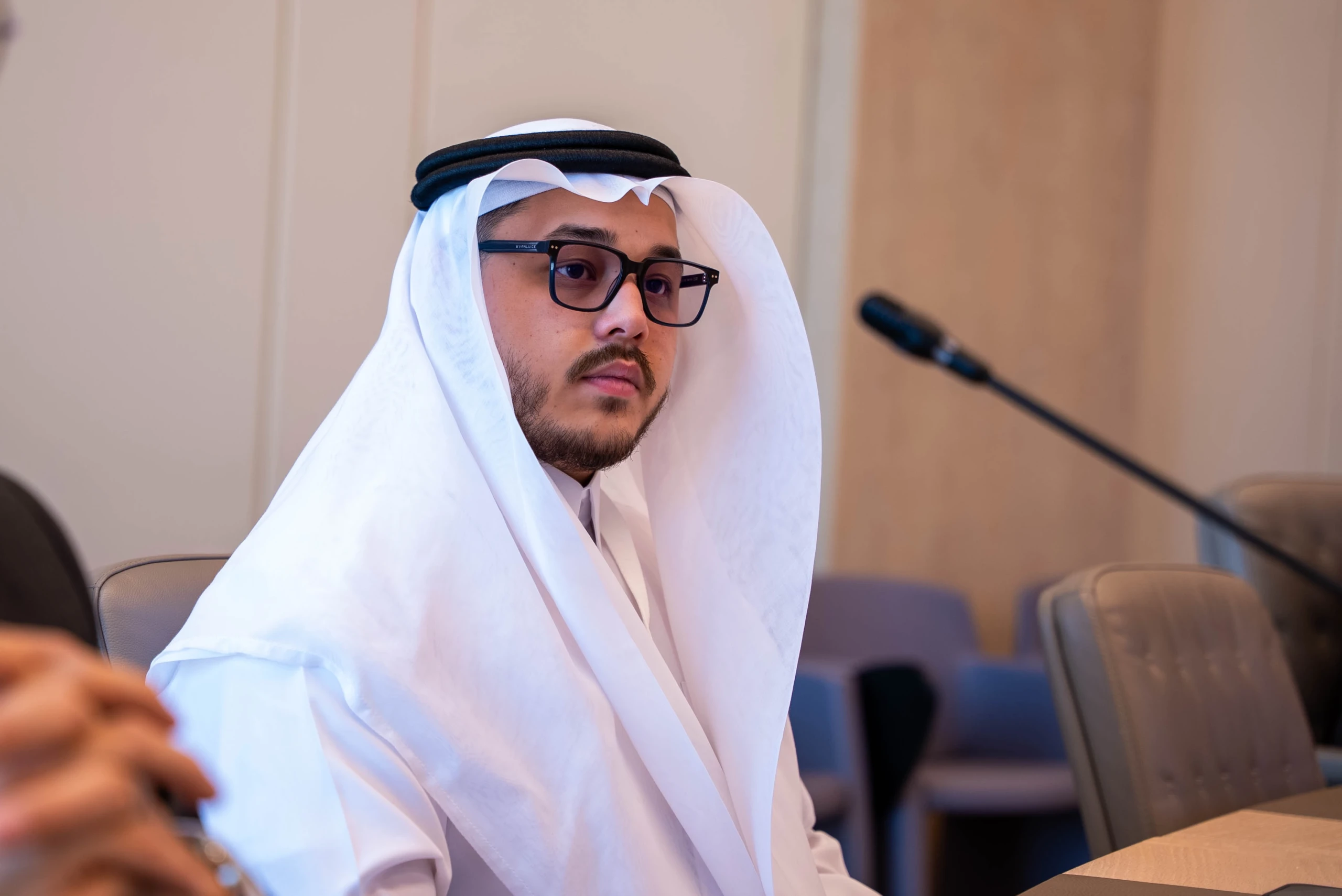 A bearded man in a white ghutra and glasses sits beside a desk microphone at a meeting.