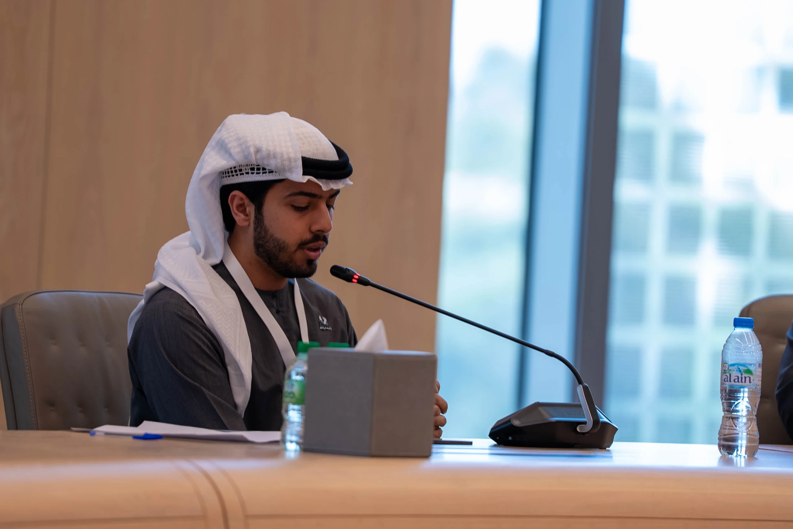 A young man in a grey kandura and white ghutra speaks into a gooseneck microphone at a table.