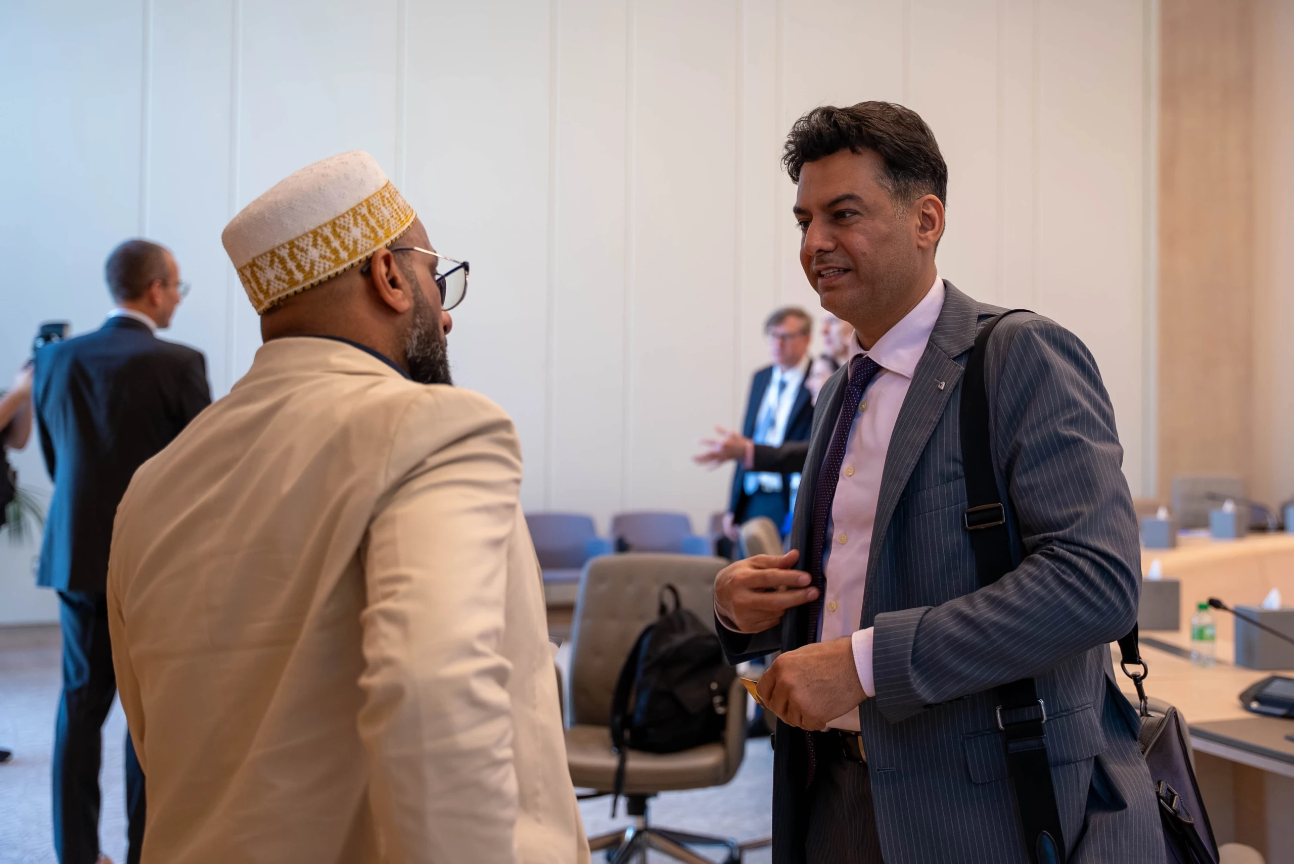 A man in a beige blazer and embroidered kufi talks with a colleague in a blue pinstripe suit.