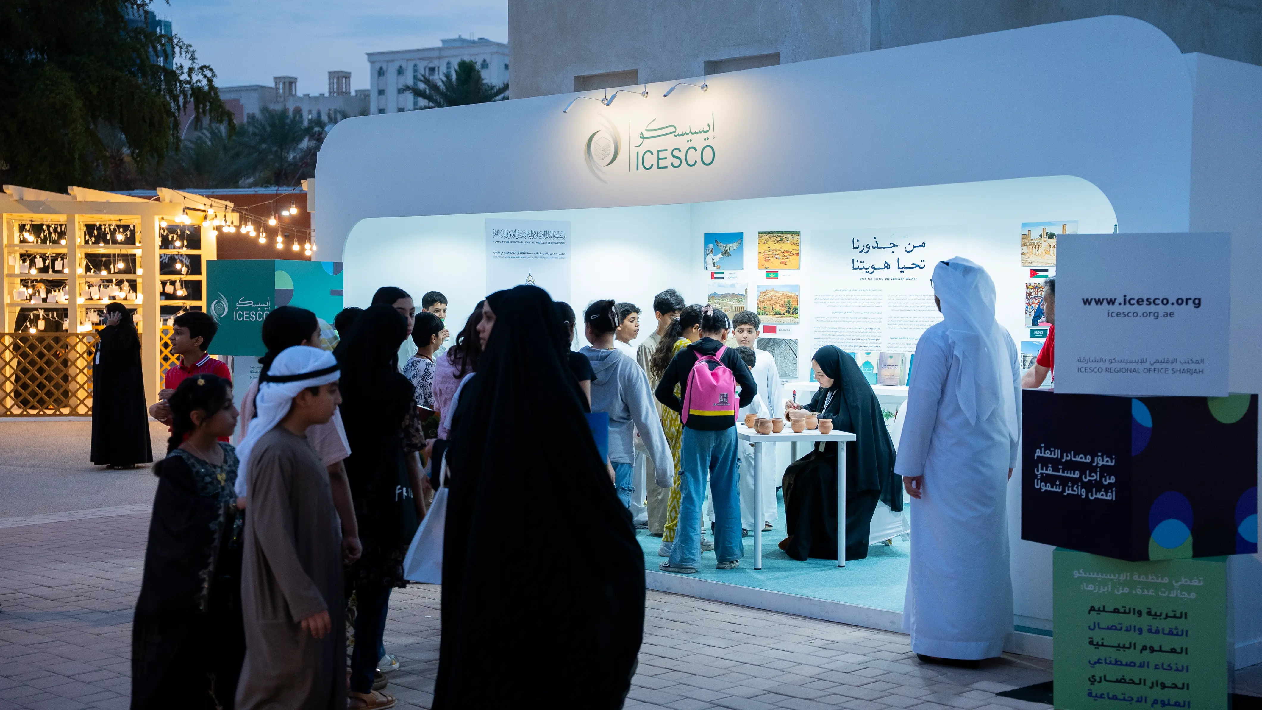 Children and visitors gather at a brightly lit ICESCO Regional Office Sharjah booth at dusk.