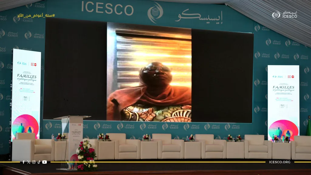 A woman in hijab appears on the central screen above empty white chairs at the ICESCO families conference.