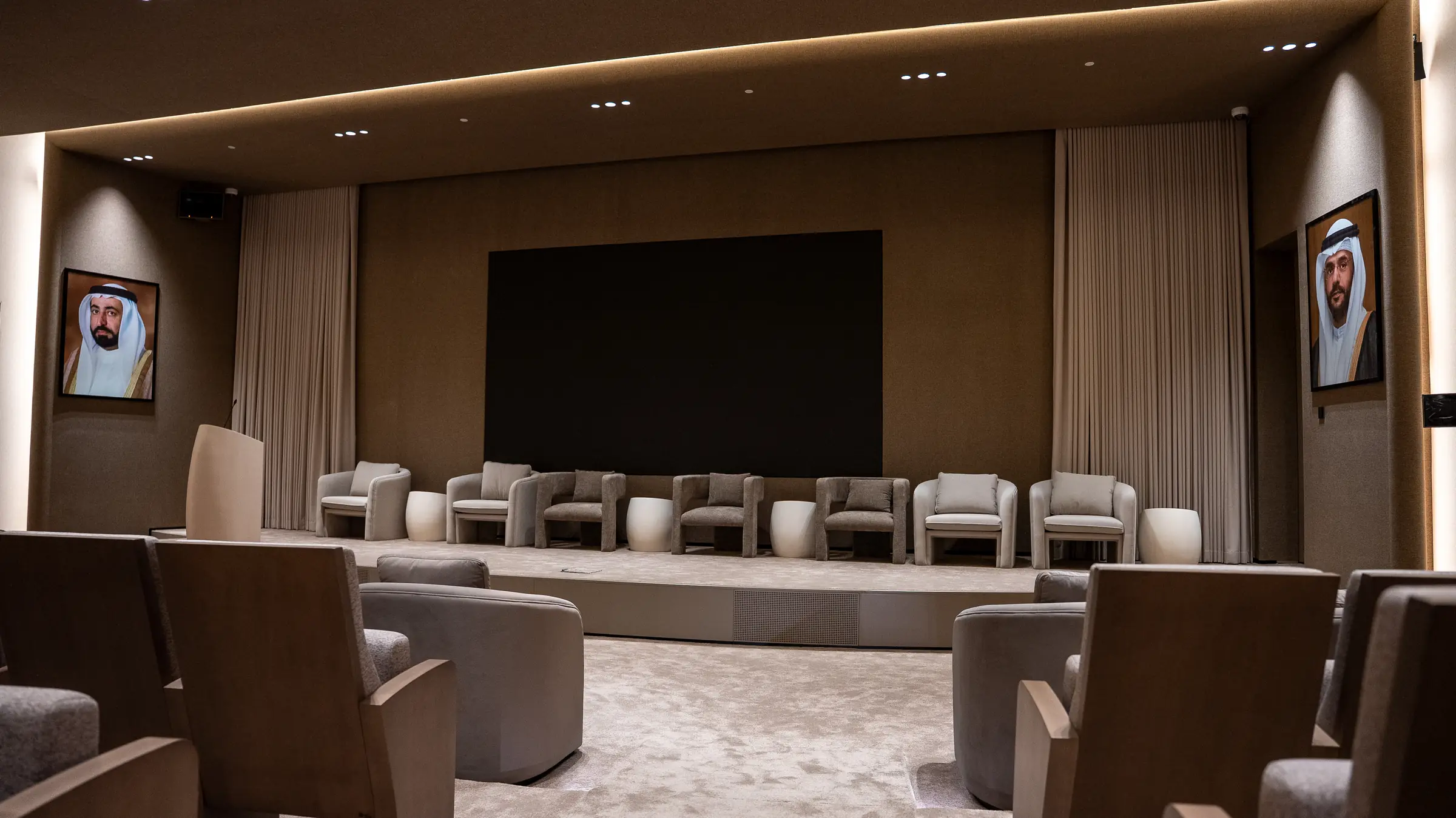 An empty conference hall with panel chairs, a wooden podium, and framed portraits of UAE leaders on the walls.