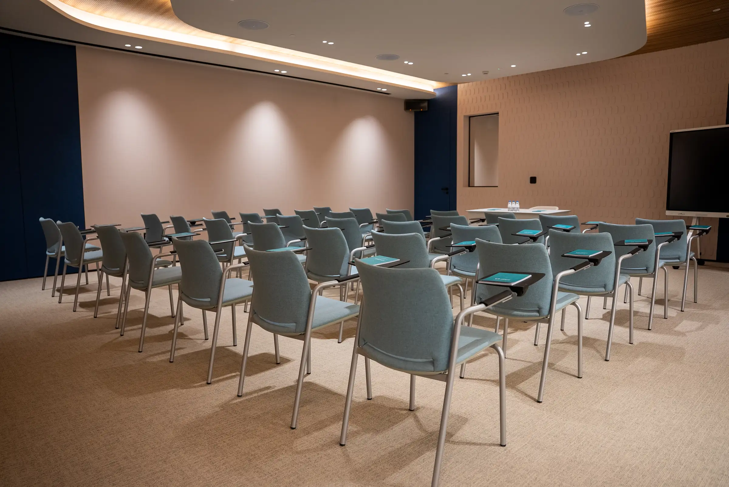 Rows of light-blue tablet-arm chairs face a front table and monitor in an empty modern training room.