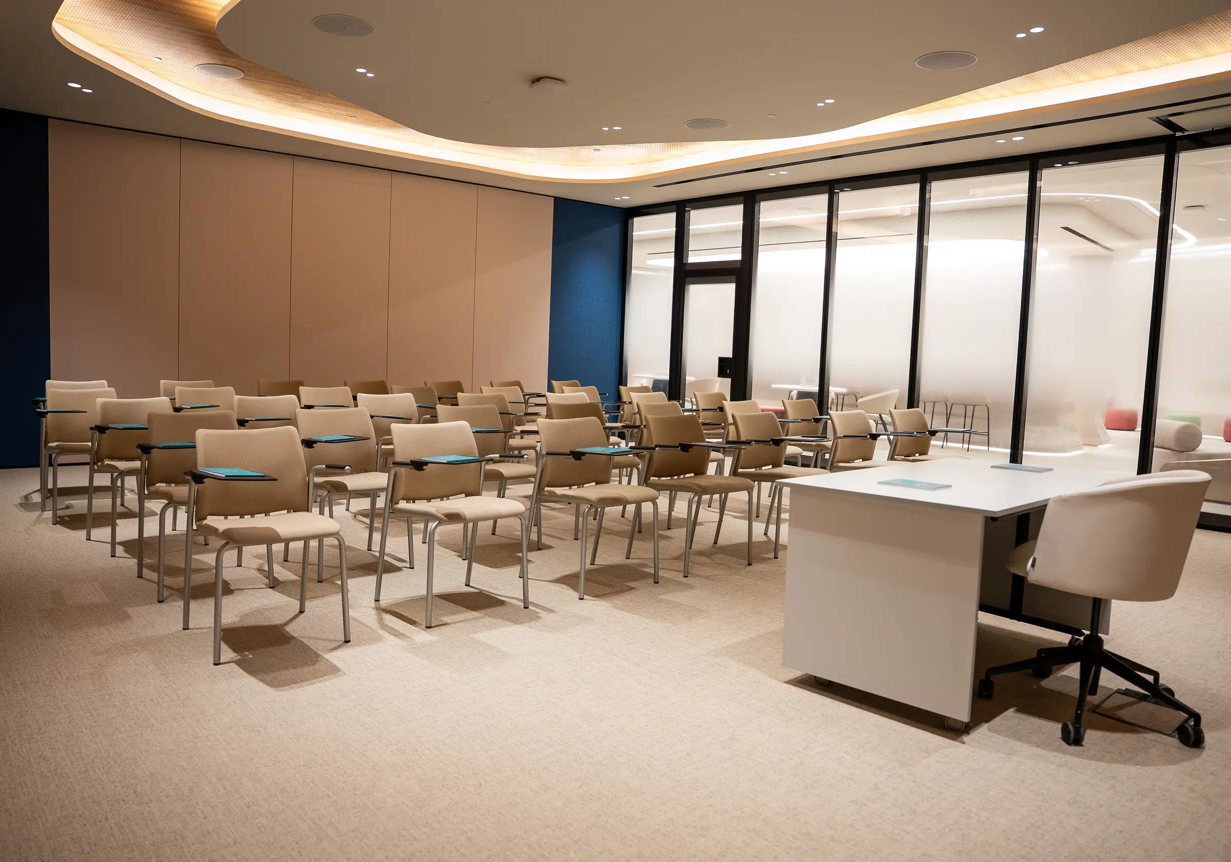 Beige tablet-arm chairs and a white presenter desk fill an empty classroom beside a glass-walled hallway.