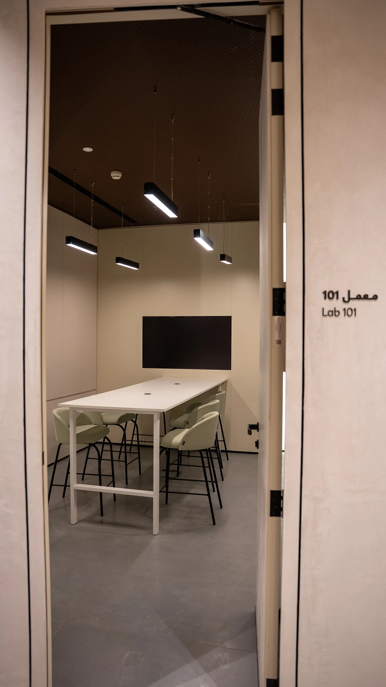 Through an open doorway labeled Lab 101, a white high table with mint stools faces a wall-mounted screen.